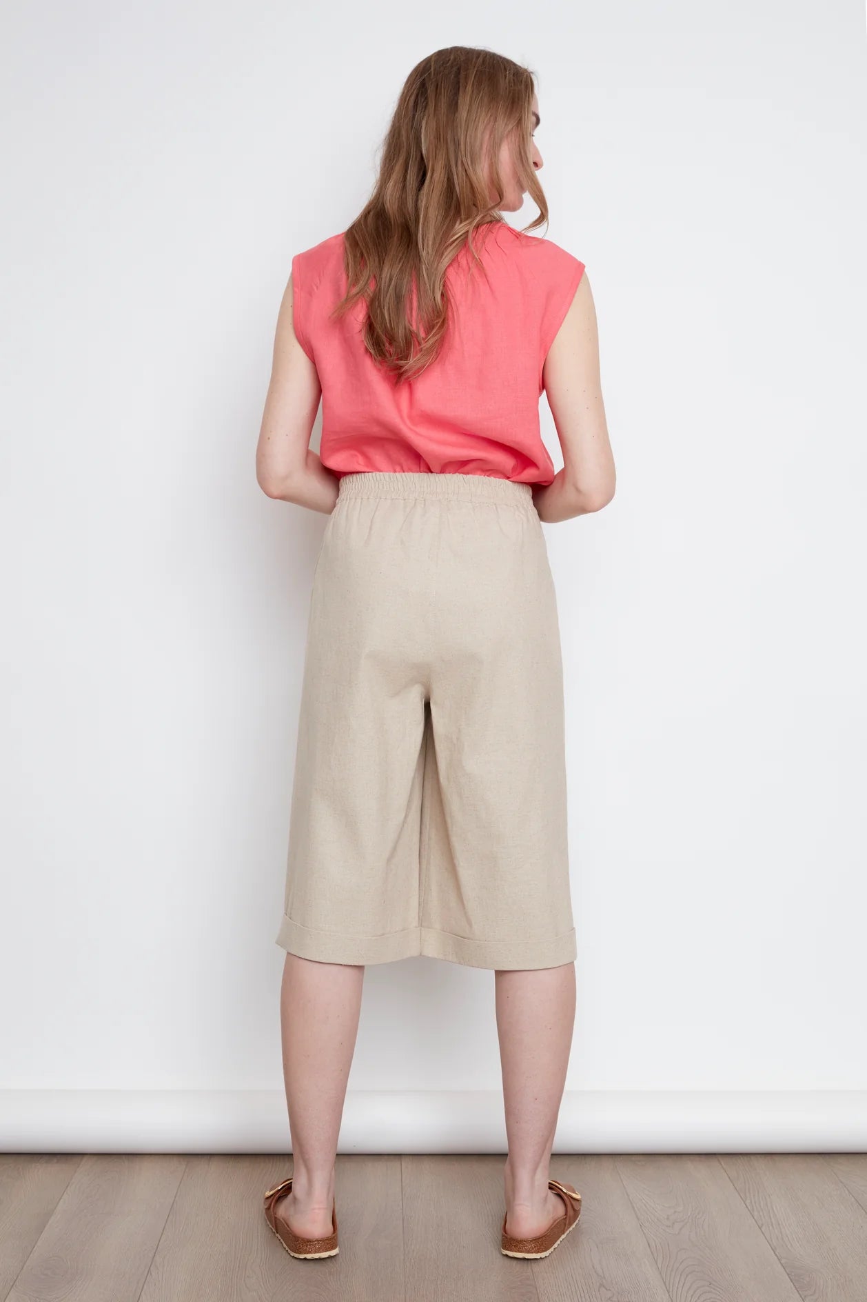 Back view of a woman wearing the Sabrina Top by Julei in coral, with raglan cap sleeves, a notched v-neck with pleat details, and a pleated back yoke. She is wearing it with the Isadora Capri pants in Natural and standing against a white background