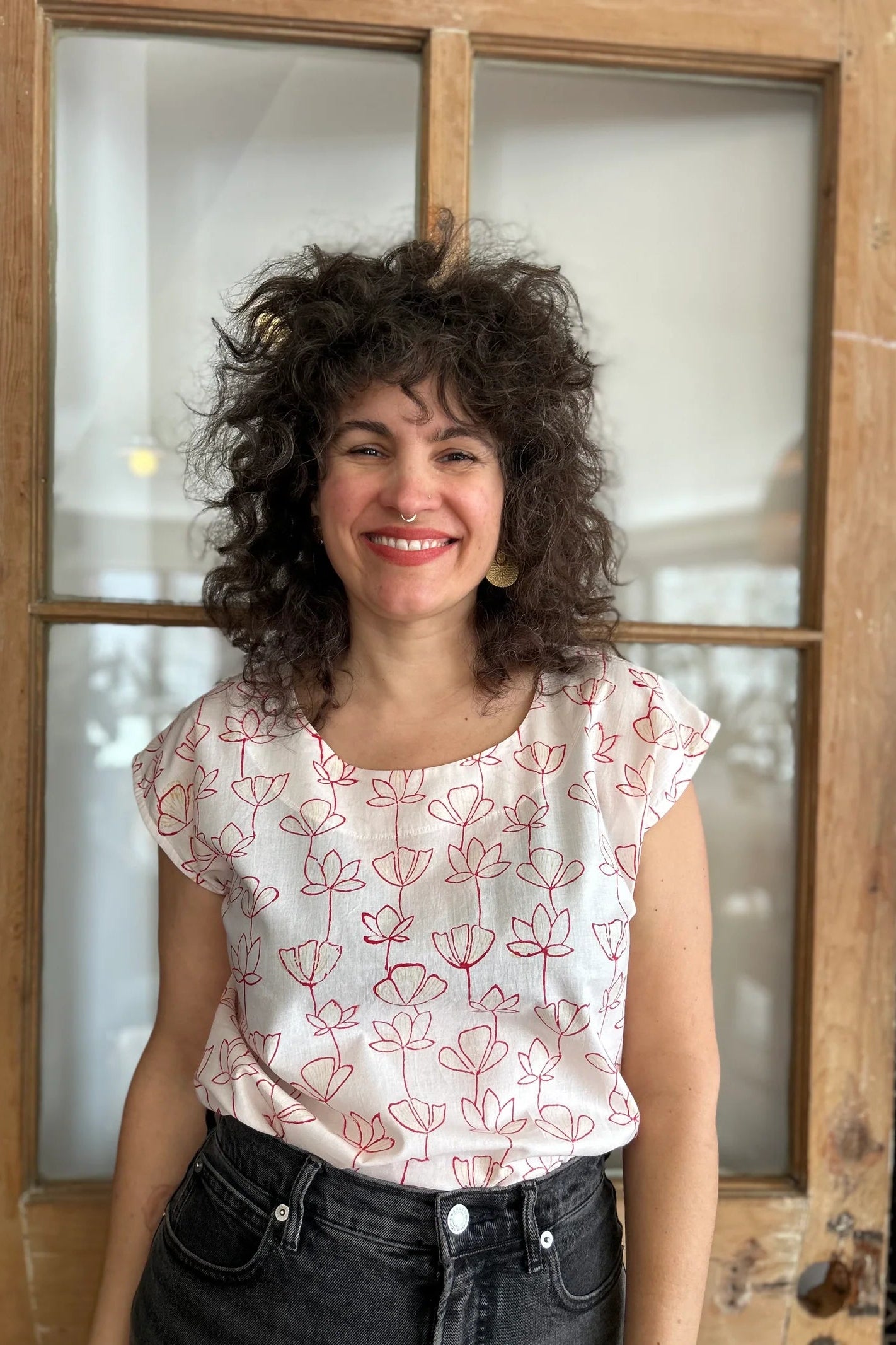 A woman wearing the wearing the Sandrigo Top from Kazak in Red Floral print, a cotton top with short extended sleeves and a wide round neck. She is wearing it black jeans and standing in front of a wooden door. 