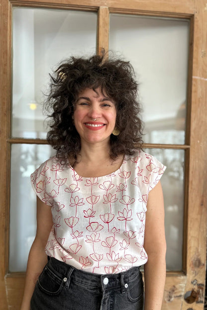 A woman wearing the wearing the Sandrigo Top from Kazak in Red Floral print, a cotton top with short extended sleeves and a wide round neck. She is wearing it black jeans and standing in front of a wooden door. 