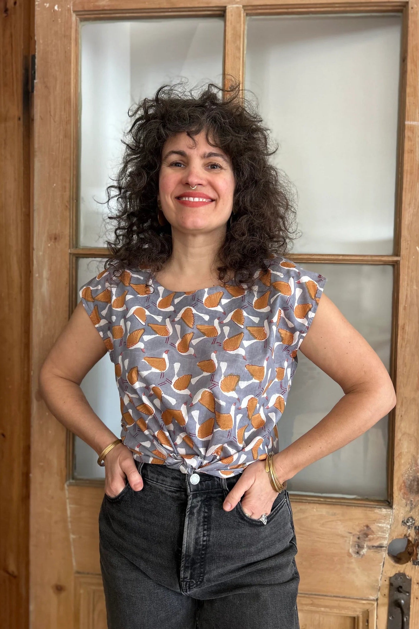 A woman wearing the wearing the Sandrigo Top from Kazak in Birds Print, a cotton top with short extended sleeves and a wide round neck. She is wearing it black jeans and standing in front of a wooden door. 