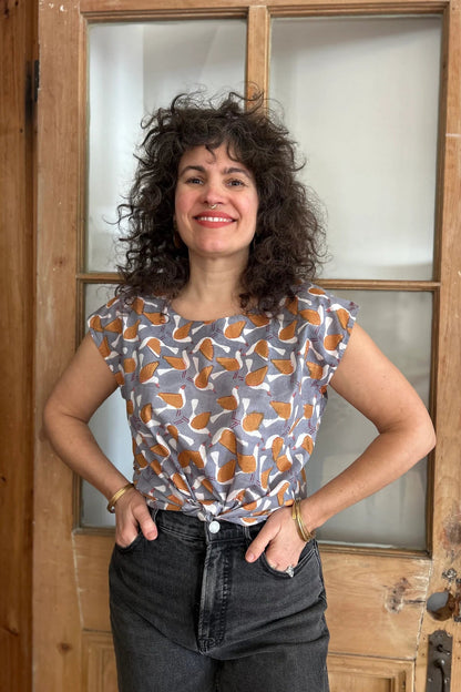 A woman wearing the wearing the Sandrigo Top from Kazak in Birds Print, a cotton top with short extended sleeves and a wide round neck. She is wearing it black jeans and standing in front of a wooden door. 