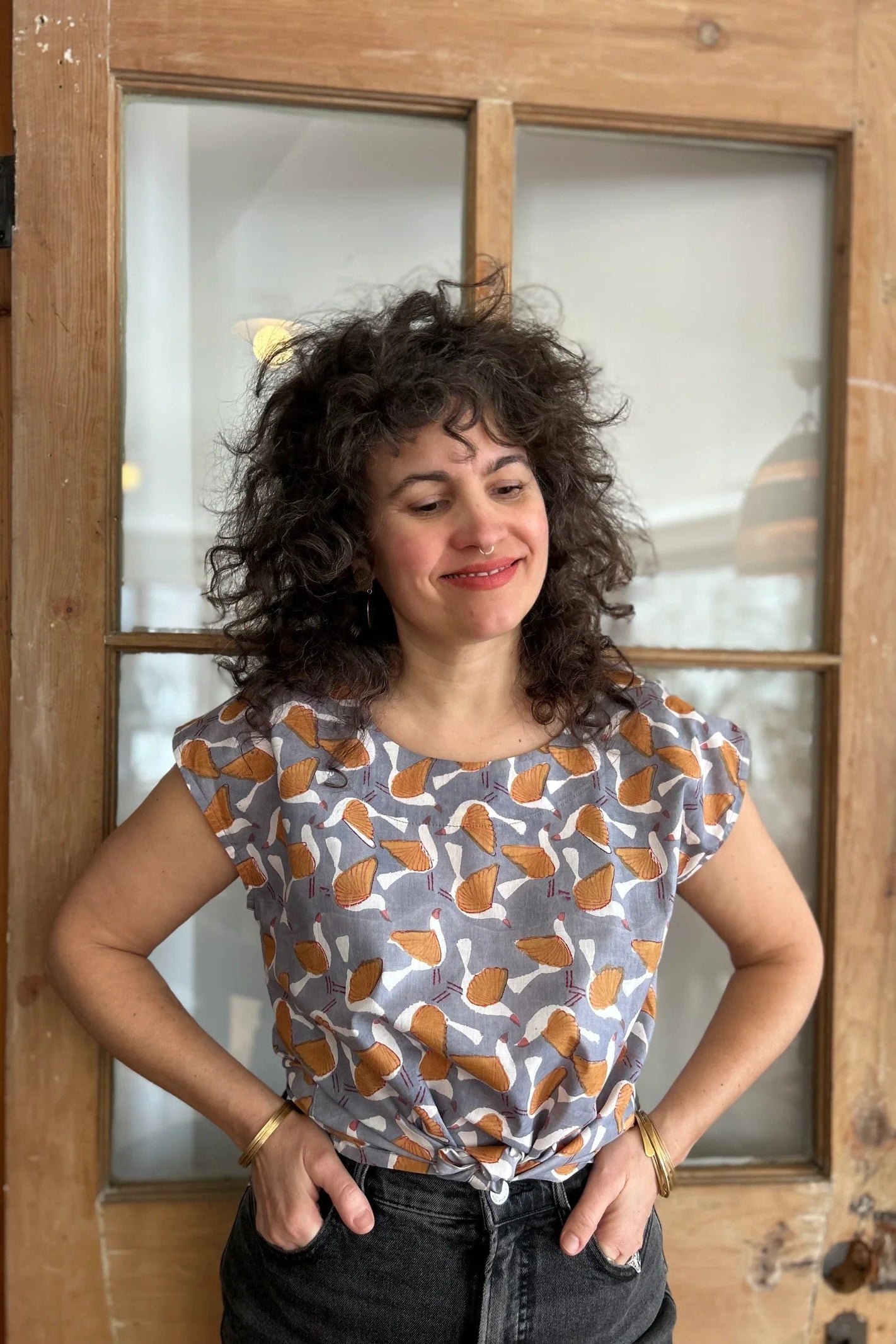 A woman wearing the wearing the Sandrigo Top from Kazak in Birds Print, a cotton top with short extended sleeves and a wide round neck. She is wearing it black jeans and standing in front of a wooden door. 