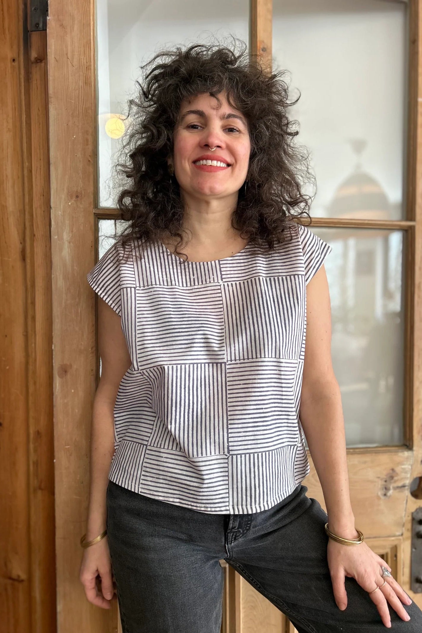 A woman wearing the wearing the Sandrigo Top from Kazak in Ori Cream print, a cotton top with short extended sleeves and a wide round neck. She is wearing it black jeans and standing in front of a wooden door. 