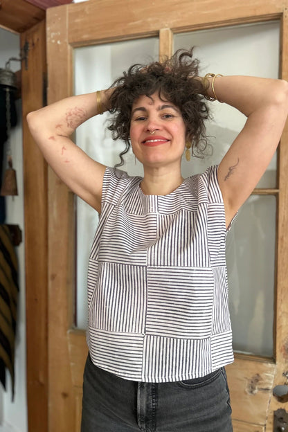 A woman wearing the wearing the Sandrigo Top from Kazak in Ori Cream print, a cotton top with short extended sleeves and a wide round neck. She is wearing it black jeans and standing in front of a wooden door. 