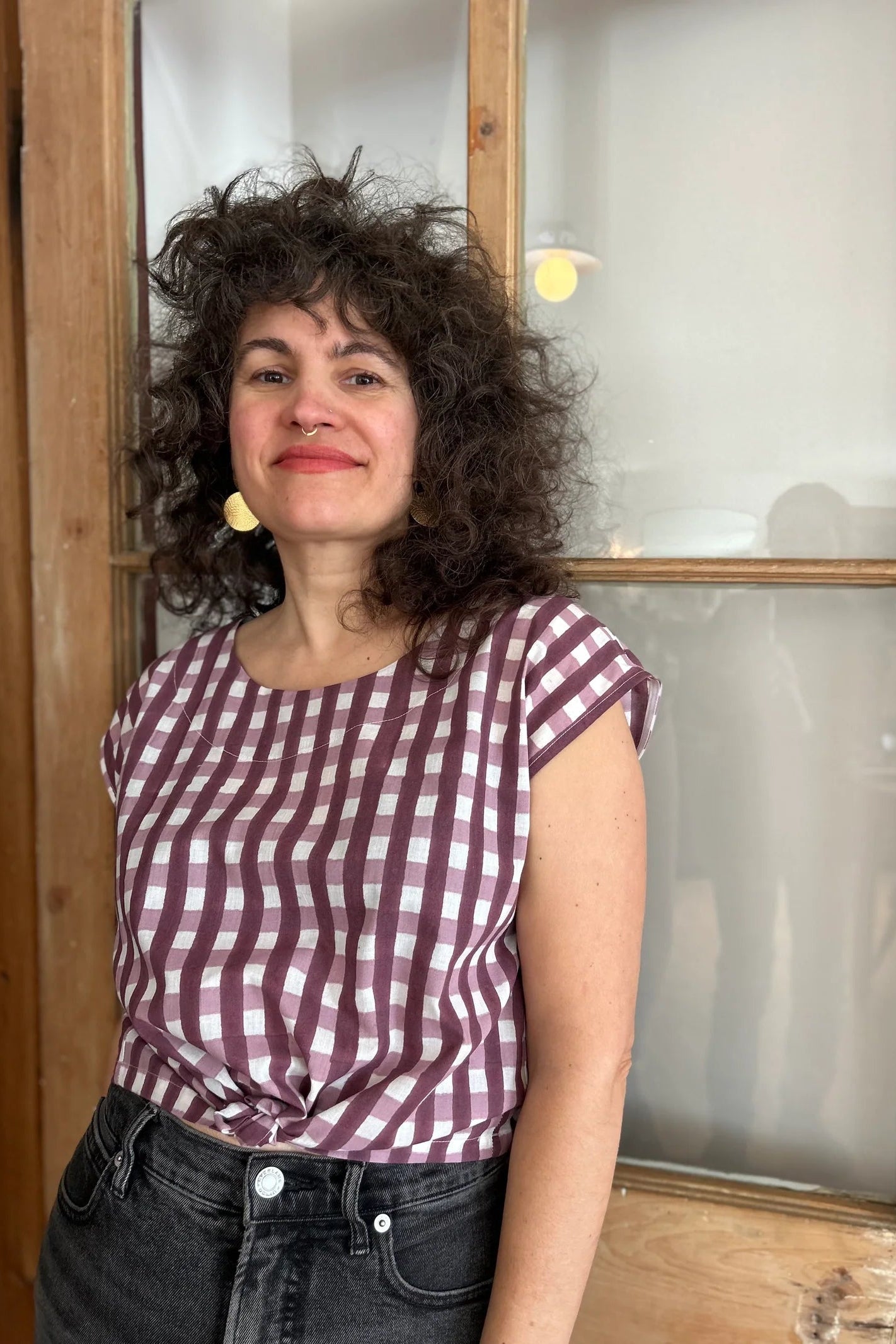 A woman wearing the wearing the Sandrigo Top from Kazak in Burgundy Gingham print, a cotton top with short extended sleeves and a wide round neck. She is wearing it black jeans and standing in front of a wooden door. 
