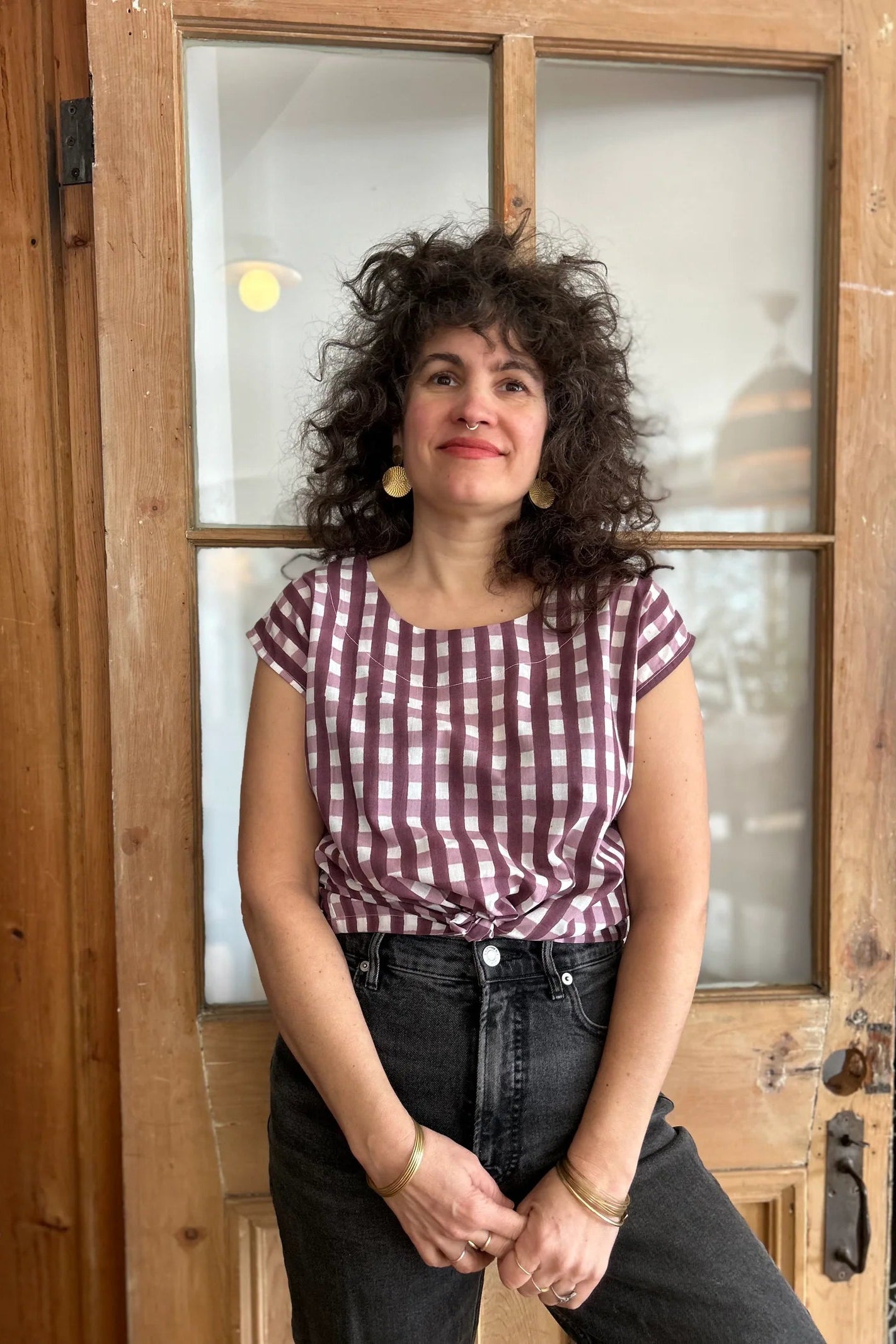 A woman wearing the wearing the Sandrigo Top from Kazak in Burgundy Gingham print, a cotton top with short extended sleeves and a wide round neck. She is wearing it black jeans and standing in front of a wooden door. 