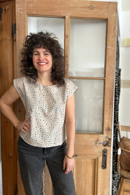 A woman wearing the wearing the Sandrigo Top from Kazak in Flowers and Insects print, a cotton top with short extended sleeves and a wide round neck. She is wearing it black jeans and standing in front of a wooden door. 