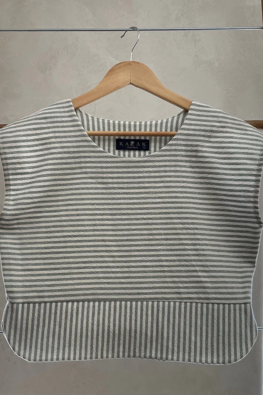 The Tulipe Top by Kazak in Sage, a short top with a round neck, short extended sleeves, horizontal stripes across the chest and a band of vertical stripes across the bottom, with a rounded hem and side slits, is shown hanging from a wooden hanger against a white wall. 