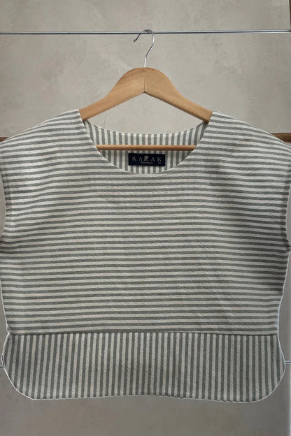The Tulipe Top by Kazak in Sage, a short top with a round neck, short extended sleeves, horizontal stripes across the chest and a band of vertical stripes across the bottom, with a rounded hem and side slits, is shown hanging from a wooden hanger against a white wall. 