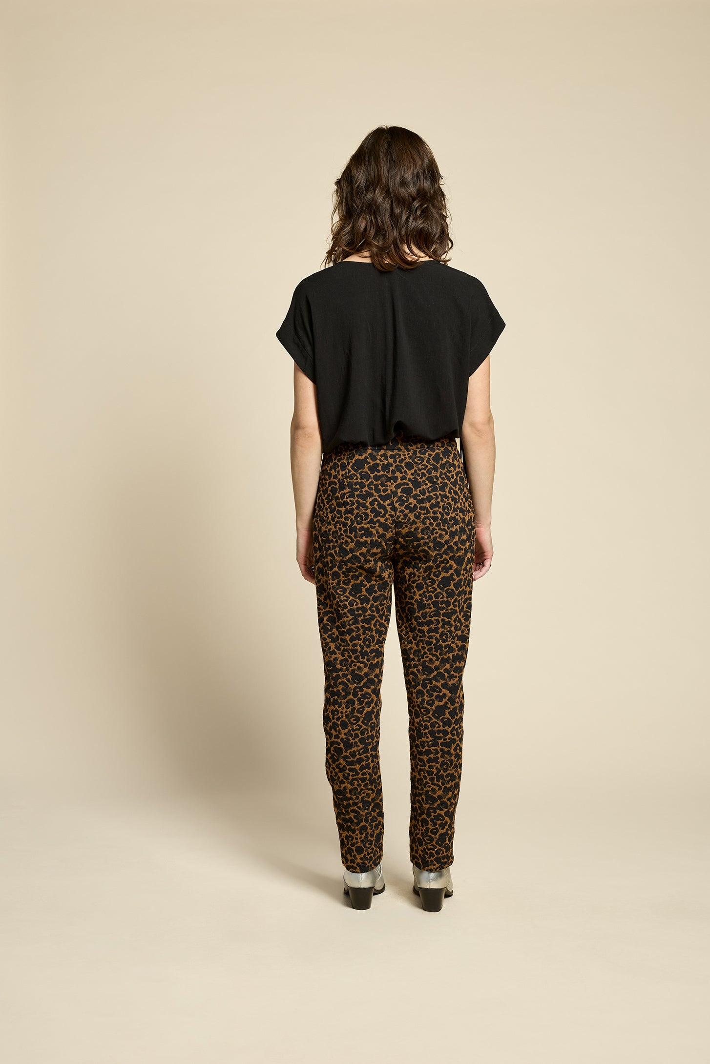 Back view of a woman wearing the Shout Top by Cokluch in Harper Black, with fringe at the shoulders, short extended sleeves, and a V-neck. She is wearing it with the Breeze Pants in Cappucino Leopard print and standing against on a beige background. 
