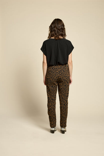 Back view of a woman wearing the Shout Top by Cokluch in Harper Black, with fringe at the shoulders, short extended sleeves, and a V-neck. She is wearing it with the Breeze Pants in Cappucino Leopard print and standing against on a beige background. 