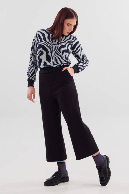 A woman wearing A24 Pants by Slak in Black, featuring a pull-on waist and wide cropped legs, with the Wanda Sweater in Blue Zebra, standing in front of a white background 