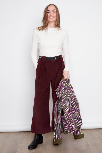 A woman wearing the Melody Mockneck Top by Julie in Natural, a ribbed knit top with a short cross-over collar and long sleeves. She is wearing it with the Isadora Pants in Burgundy corduroy and holding a patterned jacket against a white background.