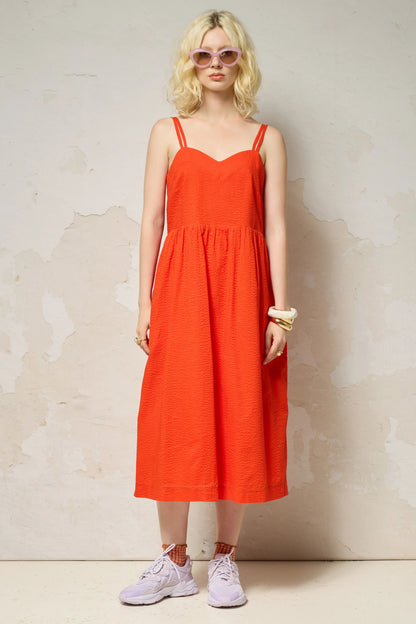 A woman wearing the Thalassa Dress by Cokluch in Fiesta, a midi dress with double spaghetti straps, a sweetheart neckline, and elastic smocking at the back, all in waffle textured cotton. She is standing against a plain background