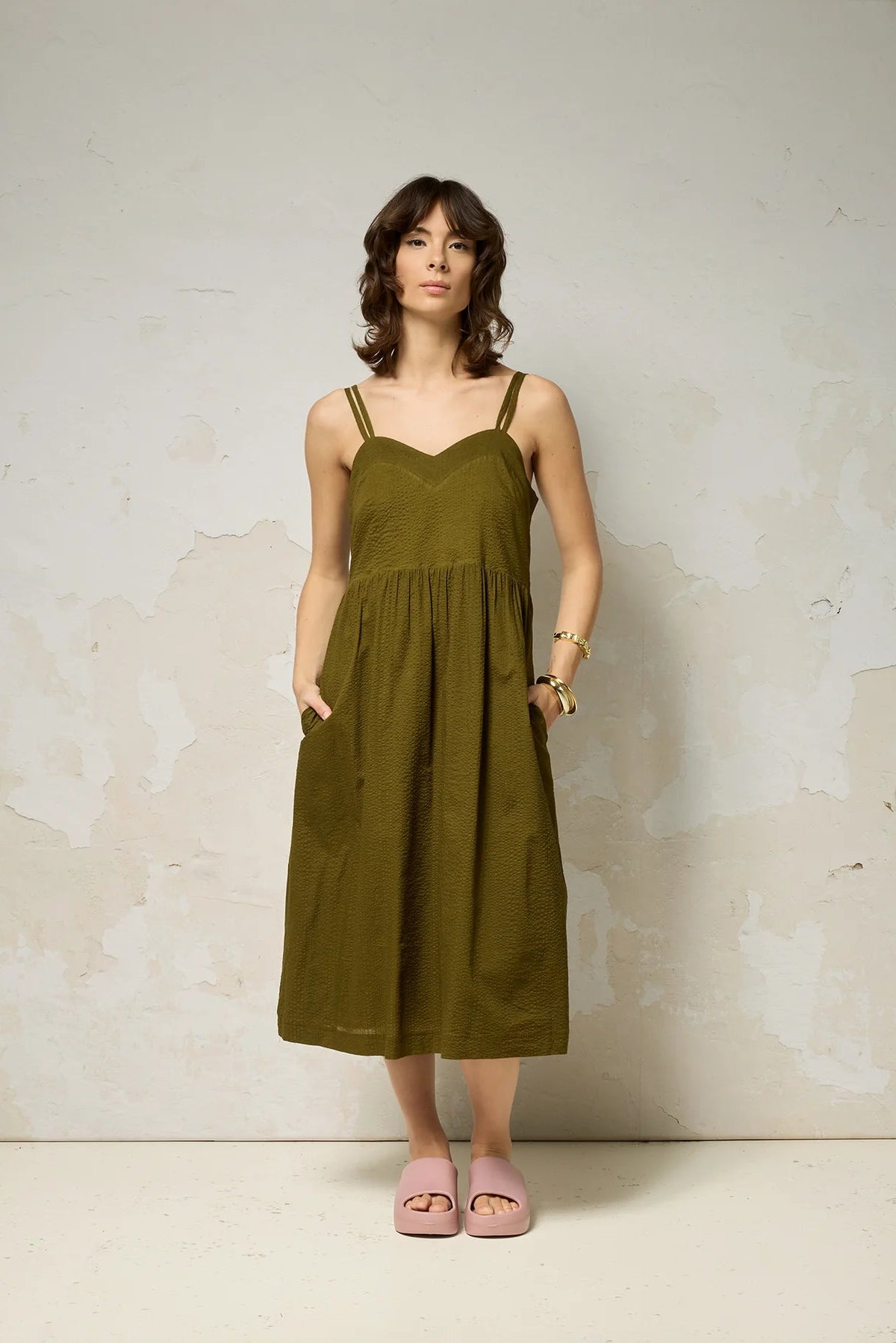 A woman wearing the Thalassa Dress by Cokluch in Olive, a midi dress with double spaghetti straps, a sweetheart neckline, and elastic smocking at the back, all in waffle textured cotton. She is standing against a plain background