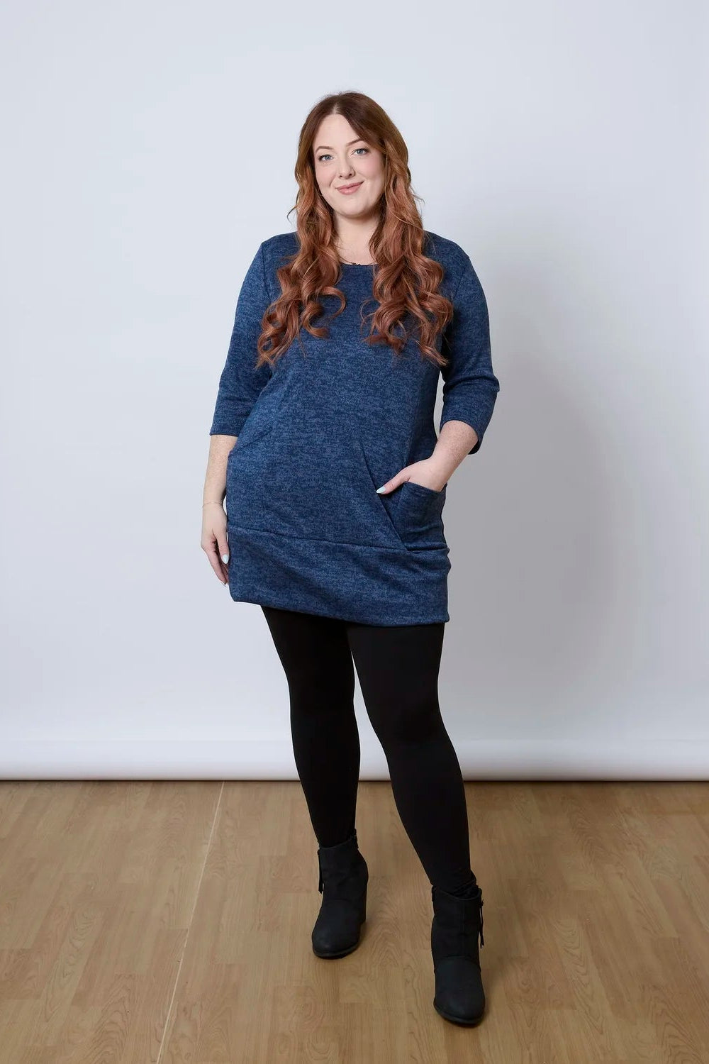 A woman wearing the Brooke Tunic by Tangente in Indigo, with 3/4 sleeves and a fitted band at the bottom. She is wearing it with black leggings and standing against a white wall. 