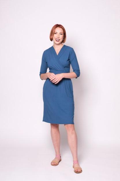 A woman wearing the Camille Dress by Tangente in Blue, featuring a cross-over neckline, narrow waistband, knee length skirt, 3/4 sleeves, and side pockets, in ribbed bamboo knit. She is standing in front of white background 