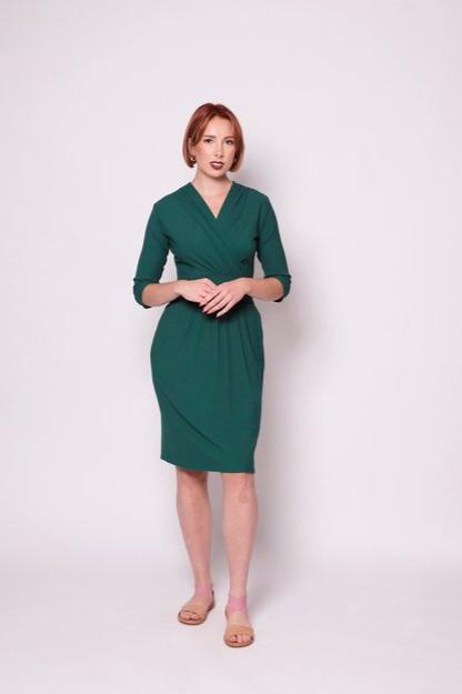 A woman wearing the Camille Dress by Tangente in Forest, featuring a cross-over neckline, narrow waistband, knee length skirt, 3/4 sleeves, and side pockets, in ribbed bamboo knit. She is standing in front of white background 