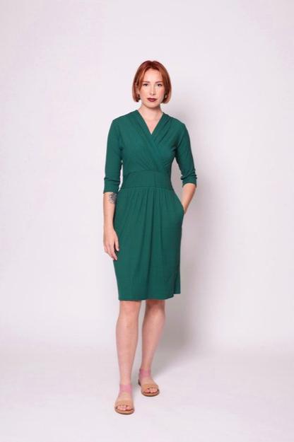 A woman wearing the Camille Dress by Tangente in Forest, featuring a cross-over neckline, narrow waistband, knee length skirt, 3/4 sleeves, and side pockets, in ribbed bamboo knit. She is standing in front of white background 
