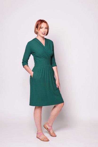 A woman wearing the Camille Dress by Tangente in Forest, featuring a cross-over neckline, narrow waistband, knee length skirt, 3/4 sleeves, and side pockets, in ribbed bamboo knit. She is standing in front of white background 