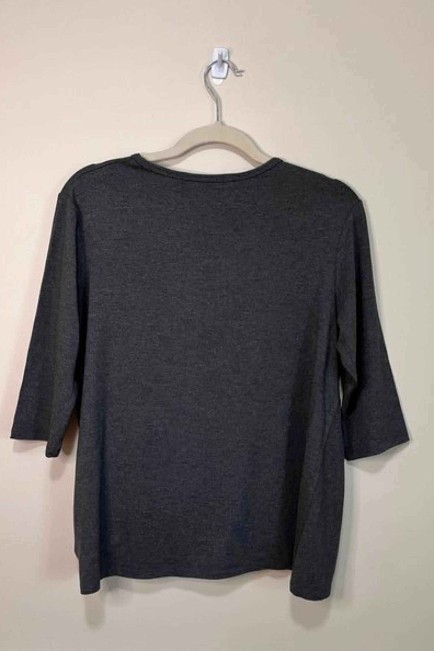 Back view of the Tangente Reese Top in Charcoal, with elbow length sleeves, a twist  at the front hem and a round neck, shown hanging in front of a beige wall 