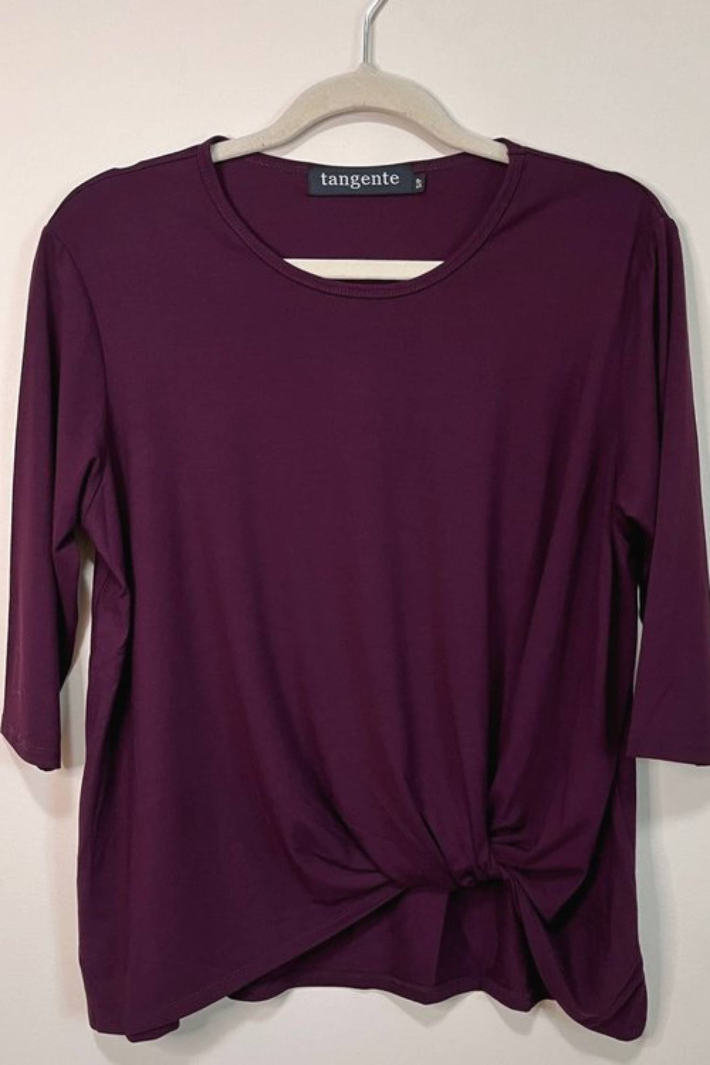 The Tangente Reese Top in Merlot, with elbow length sleeves, a twist  at the front hem and a round neck, is shown hanging in front of a beige wall 