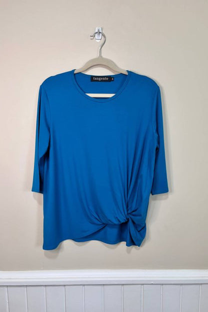 The Tangente Reese Top in Turquoise, with elbow length sleeves, a twist  at the front hem and a round neck, is shown hanging in front of a beige wall 