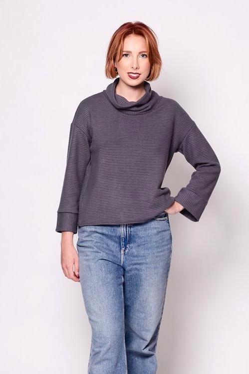 A woman wearing the Sidney Top by Tangente in Charcoal, a thick ribbed knit sweater with a cowl neck, long cuffed sleeves, and a slightly boxy fit. She's wearing it with jeans and standing in front of a white background. 