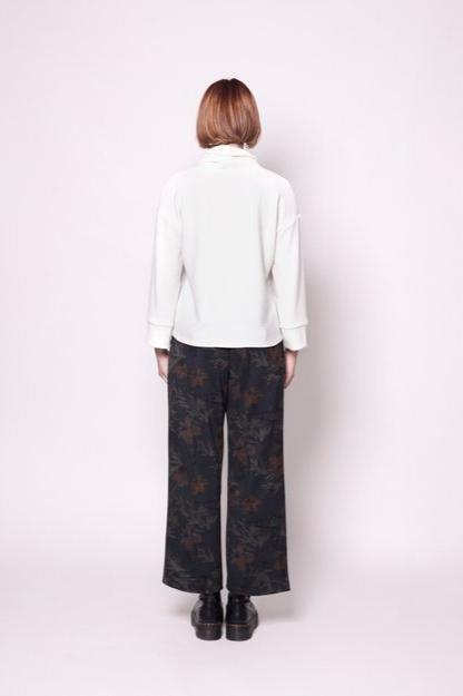 Back view of a woman wearing the Sidney Top by Tangente in Off-white, a thick ribbed knit sweater with a cowl neck, long cuffed sleeves, and a slightly boxy fit. She's wearing it with floral pants and standing in front of a white background. 