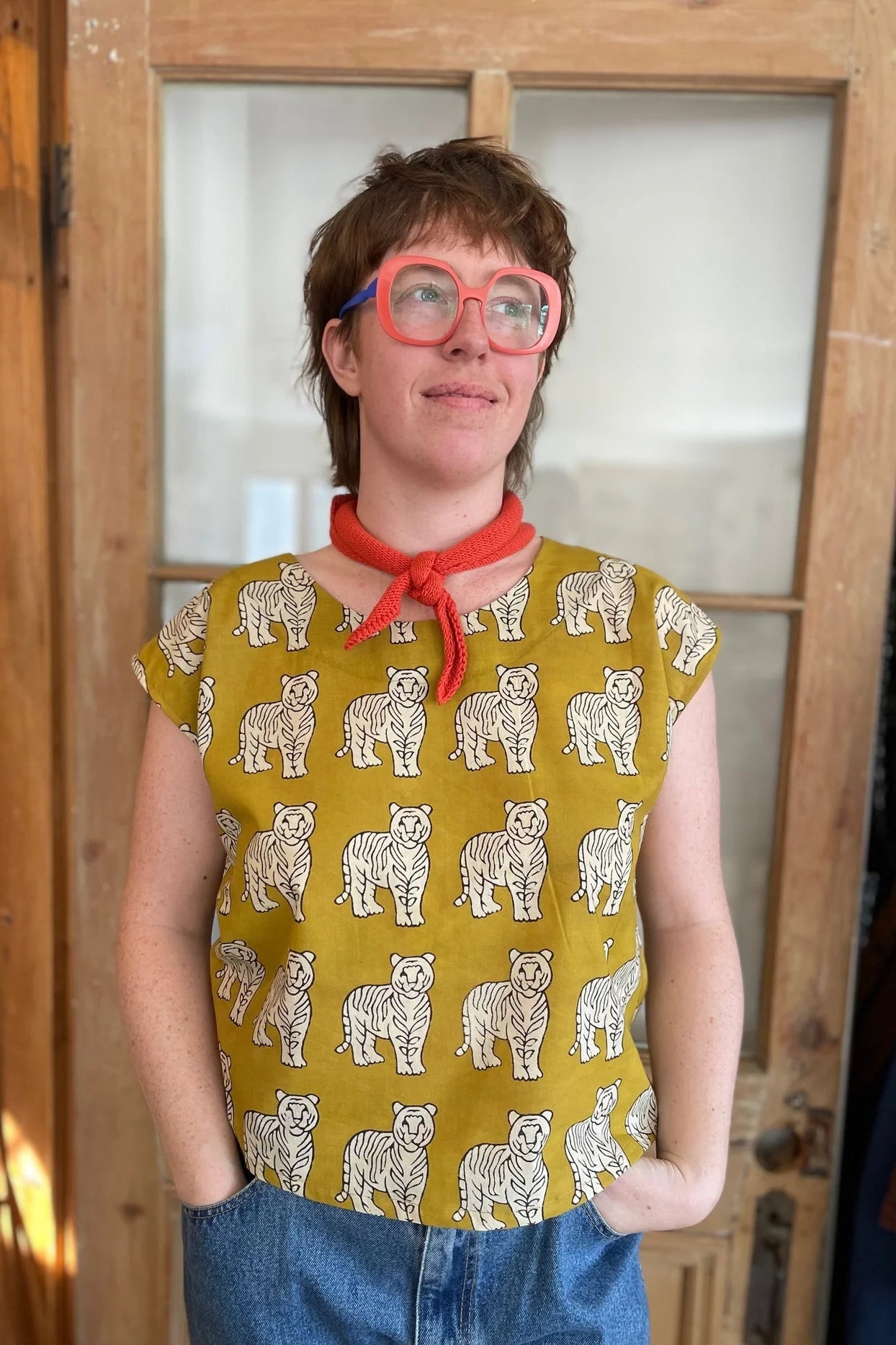 A woman wearing the wearing the Sandrigo Top from Kazak in Tigers, a loose cotton top with short extended sleeves and a wide round neck. She is wearing it light jeans and an orange scarf and standing in front of a wooden door. 