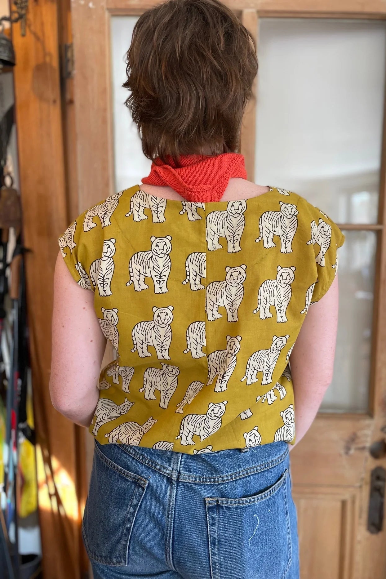 Back view of a woman wearing the wearing the Sandrigo Top from Kazak in Tigers, a loose cotton top with short extended sleeves and a wide round neck. She is wearing it light jeans and an orange scarf and standing in front of a wooden door. 