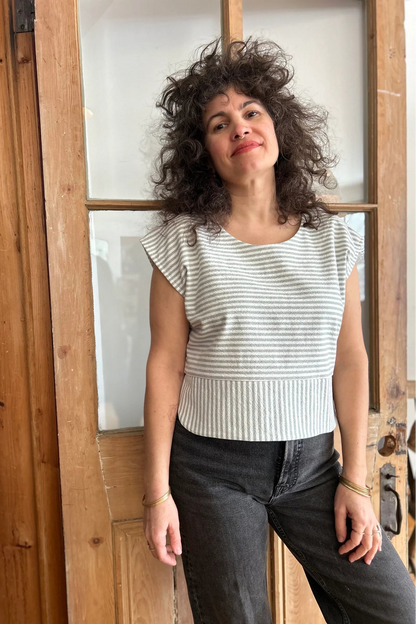 A woman wearing the Tulipe Top by Kazak in Sage, a short top with a round neck, short extended sleeves, horizontal stripes across the chest and a band of vertical stripes across the bottom, with a rounded hem and side slits. She is wearing it with black jeans and standing in front of a wooden door. 