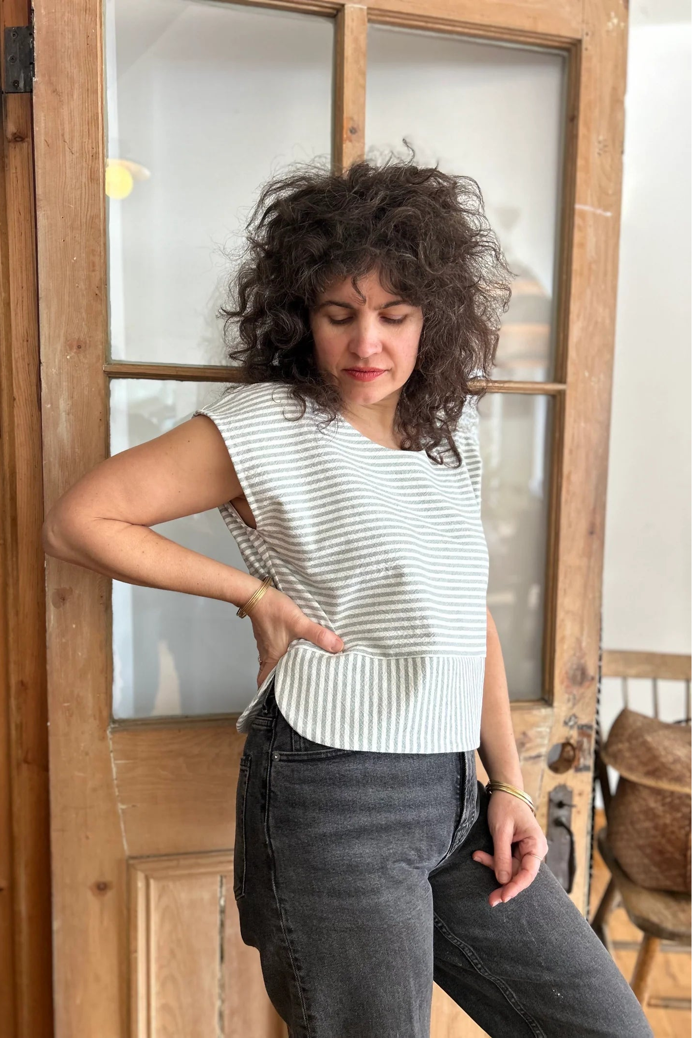 A woman wearing the Tulipe Top by Kazak in Sage, a short top with a round neck, short extended sleeves, horizontal stripes across the chest and a band of vertical stripes across the bottom, with a rounded hem and side slits. She is wearing it with black jeans and standing in front of a wooden door. 