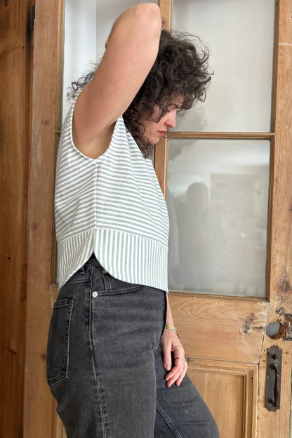 Side view of a woman wearing the Tulipe Top by Kazak in Sage, a short top with a round neck, short extended sleeves, horizontal stripes across the chest and a band of vertical stripes across the bottom, with a rounded hem and side slits. She is wearing it with black jeans and standing in front of a wooden door. 