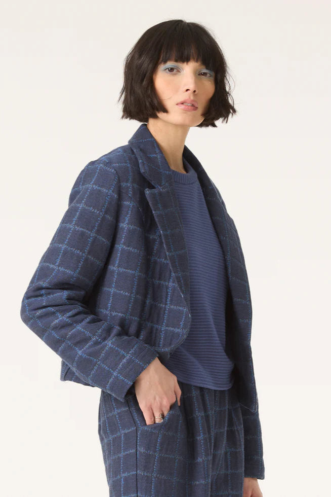 3/4 view of a woman wearing the Unreal Jacket by Cokluch in Denim, featuring a subtle plaid pattern, an open tailored collar, a short and slightly loose fit, and a two button closure. She is wearing with with a blue top and the matching Vega pants and is standing in front of a white background. 