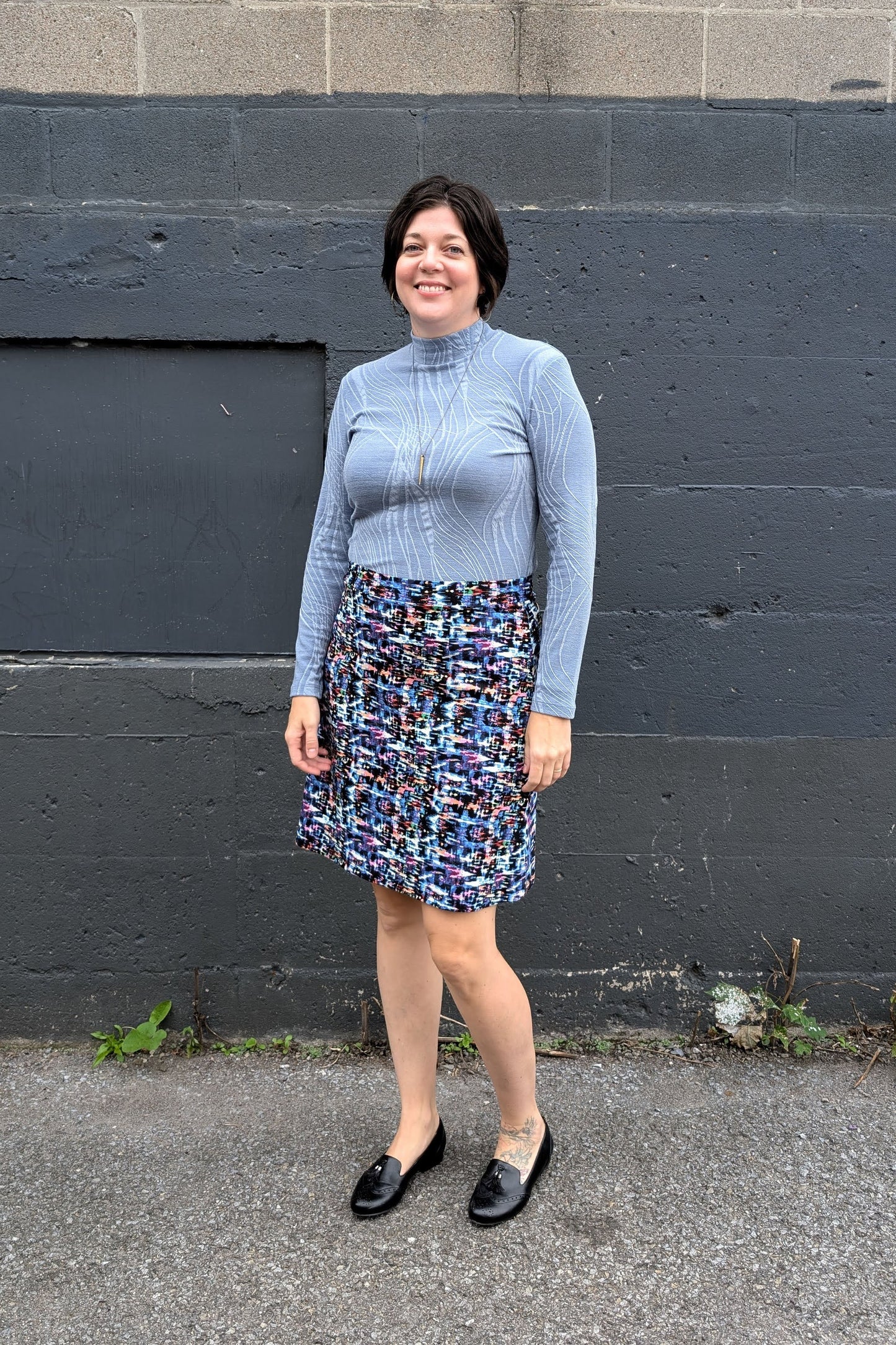 A woman wearing the Franco Mock Top by Luc Fontaine in blue, featuring a textured fabric, mock turtleneck and long sleeves, with the Eve Skirt by Slak, standing outside in front of a brick wall. 