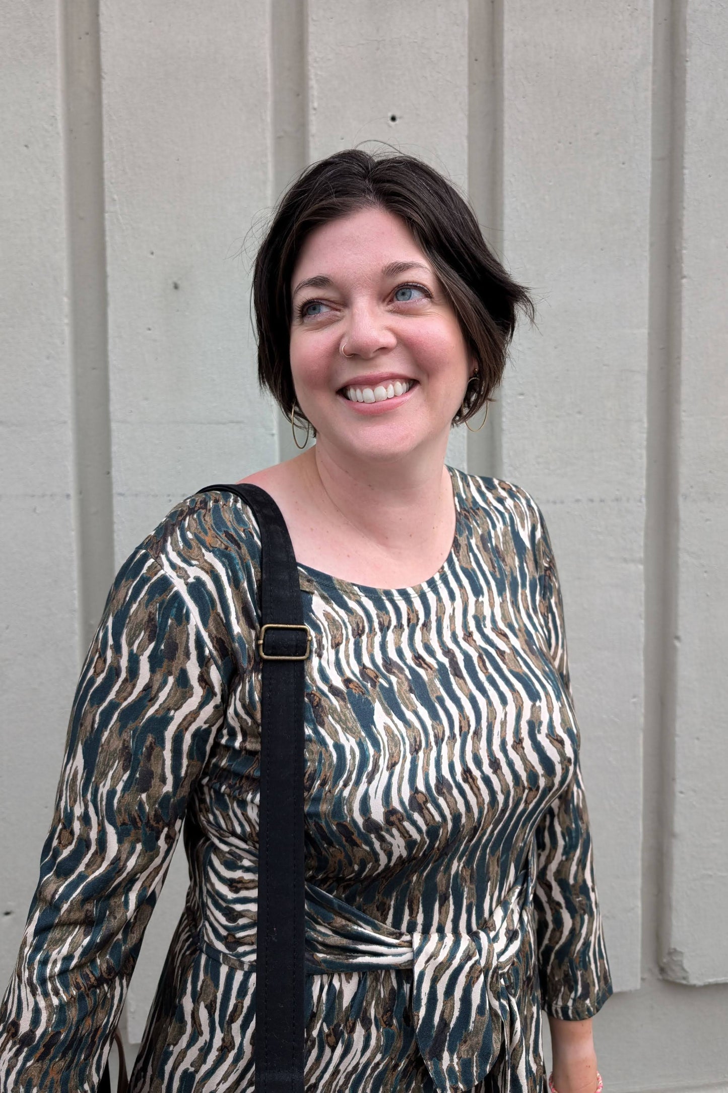 Close up of a woman wearing the Gwen Dress by Tangente in Green print, standing outside in front of a white wall
