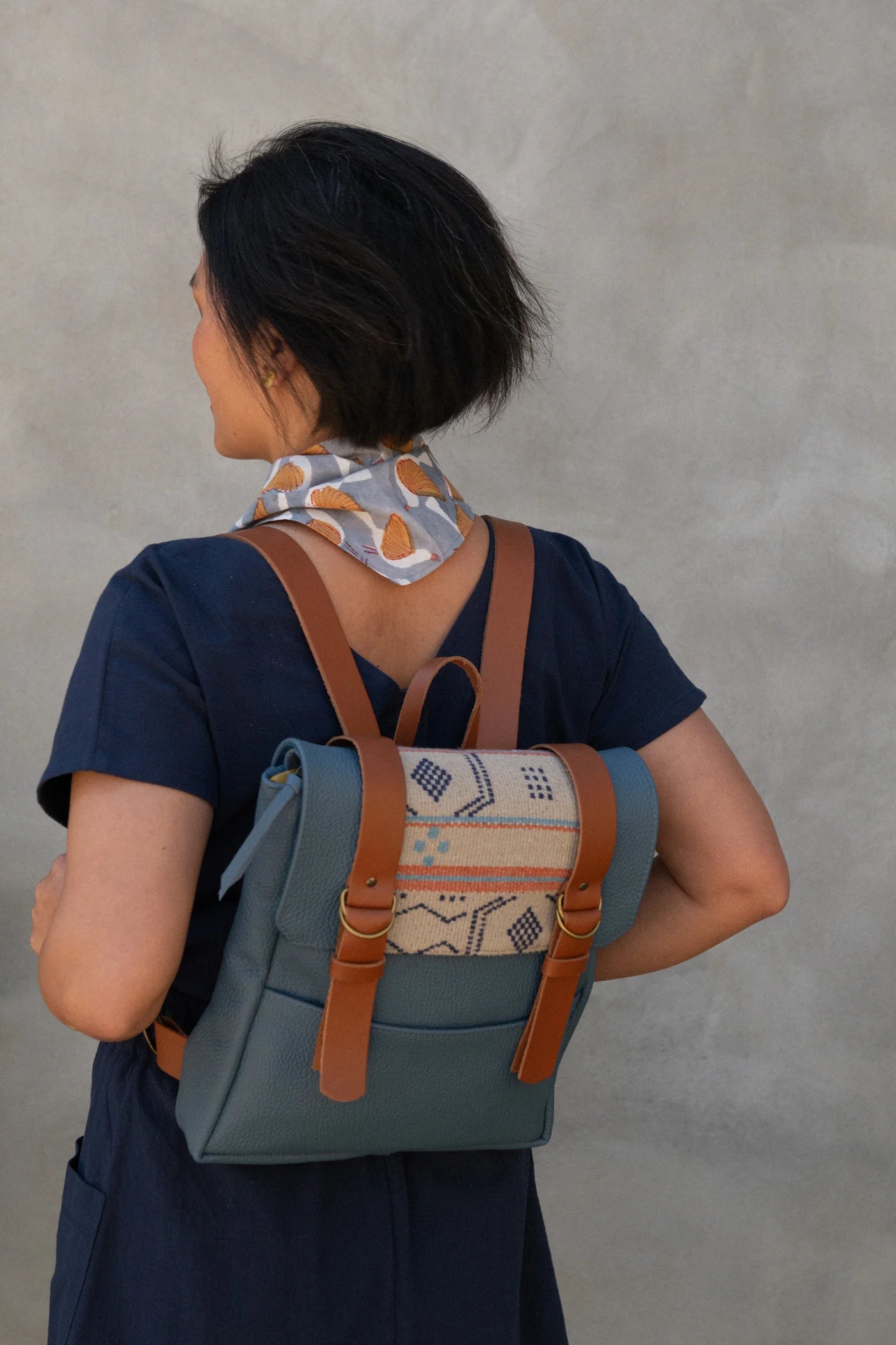 Back view of a woman wearing the Brendola Top from Kazak in Navy, a slightly cropped short sleeved top with three brass snaps that can be worn at the back or the front. She is wearing it with a scarf and the Nymphea skirt in Navy and a Kazak backpack, and standing outside in front of a grey wall.