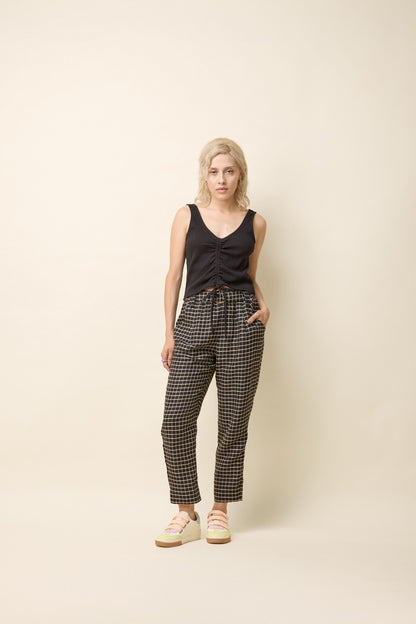 A woman wearing the Yasmina Pants from Cokluch in Black Utopia, made from checkered linen and cotton with a high elastic waist with a ring belt, and tapered, ankle-length legs. She is wearing them with a black top and standing in front of a white background. 