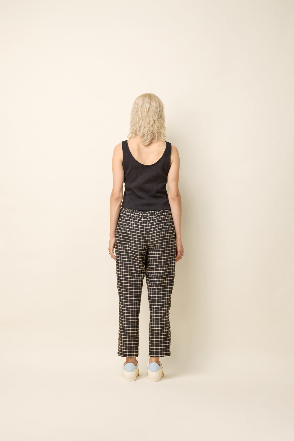 Back view of a woman wearing the Yasmina Pants from Cokluch in Black Utopia, made from checkered linen and cotton with a high elastic waist with a ring belt, and tapered, ankle-length legs. She is wearing them with a black top and standing in front of a white background. 