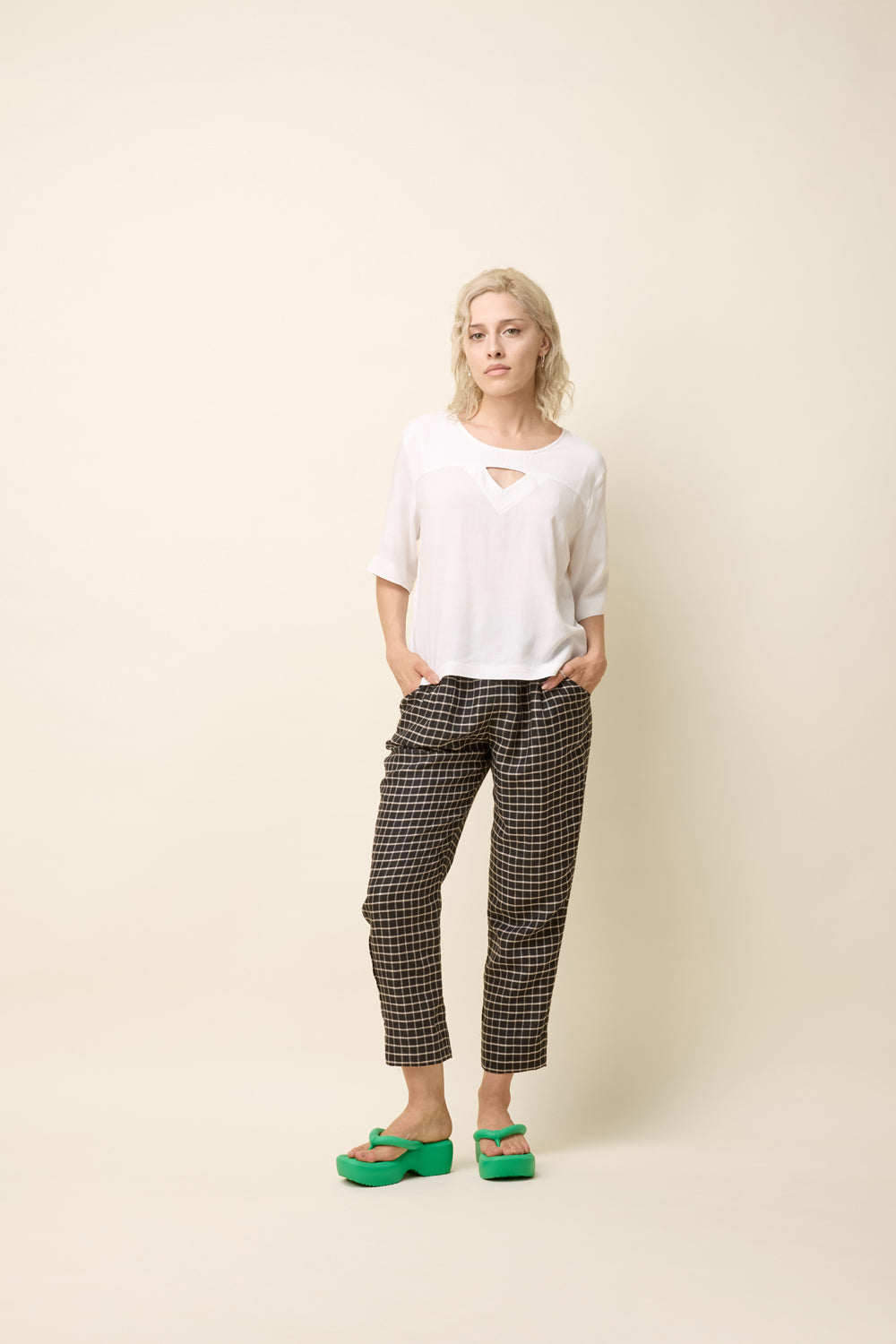 A woman wearing the Yasmina Pants from Cokluch in Black Utopia, made from checkered linen and cotton with a high elastic waist with a ring belt, and tapered, ankle-length legs. She is wearing them with a white top and standing in front of a beige background. 