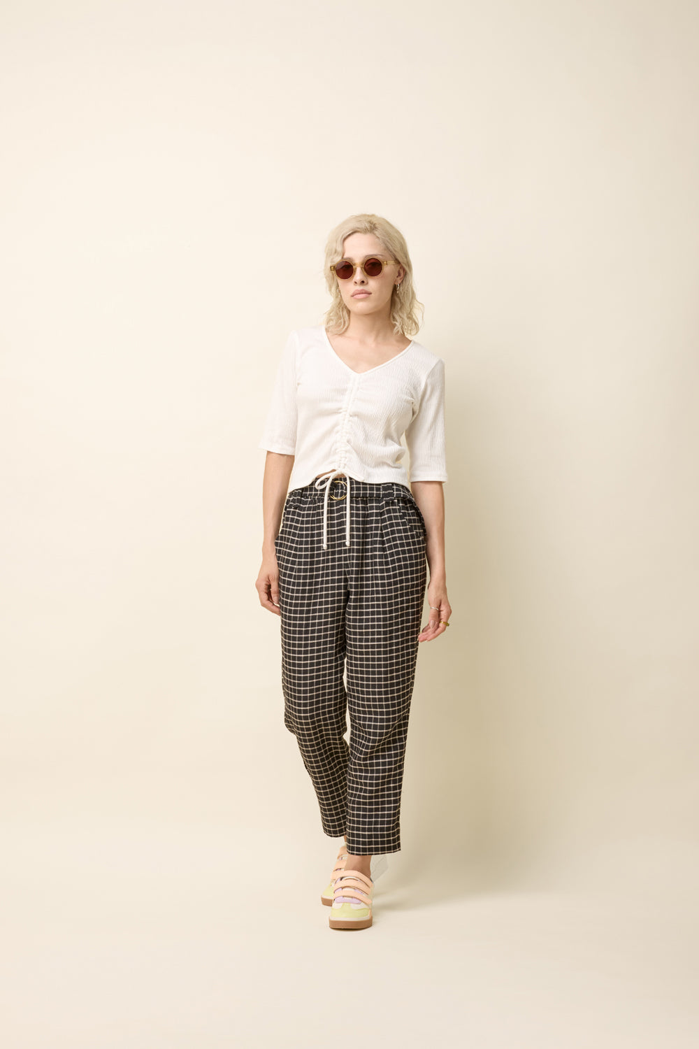 A woman wearing the Yasmina Pants from Cokluch in Black Utopia, made from checkered linen and cotton with a high elastic waist with a ring belt, and tapered, ankle-length legs. She is wearing them with a white top and standing in front of a beige background. 