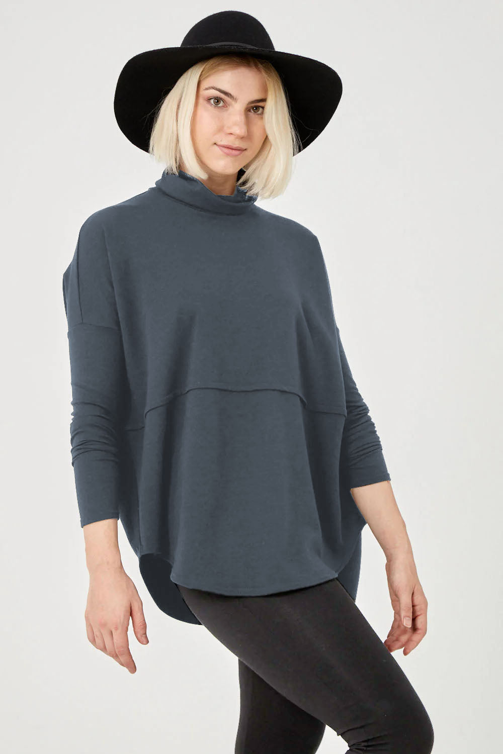 A woman wearing the Abigail Tunic by Advika in Steel, featuring a turtleneck, long extended sleeves, a seam across the body, and a rounded hem, in cotton/tencel fabric. She is wearing it with leggings and standing in front of a white background. 