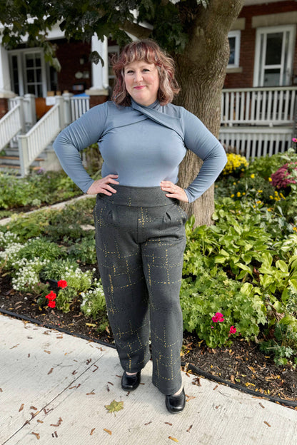 A woman wearing the Jacques Pants by Melow in Anthracite with a faint mustard check, featuring a high pull-on waistband, front pleats, and straight legs, with the Julei Irina Sweater in Ocean. She is standing outside in front of a garden. 