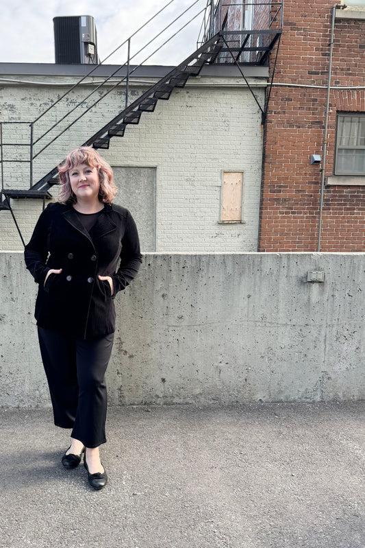 A woman wearing the Mission Blazer by Luc Fontaine in Black, in black corduroy that's slightly sheer between the wales, with a double-breasted front and front pockets. She is wearing it with black pants and standing outside in front of a brick wall. 