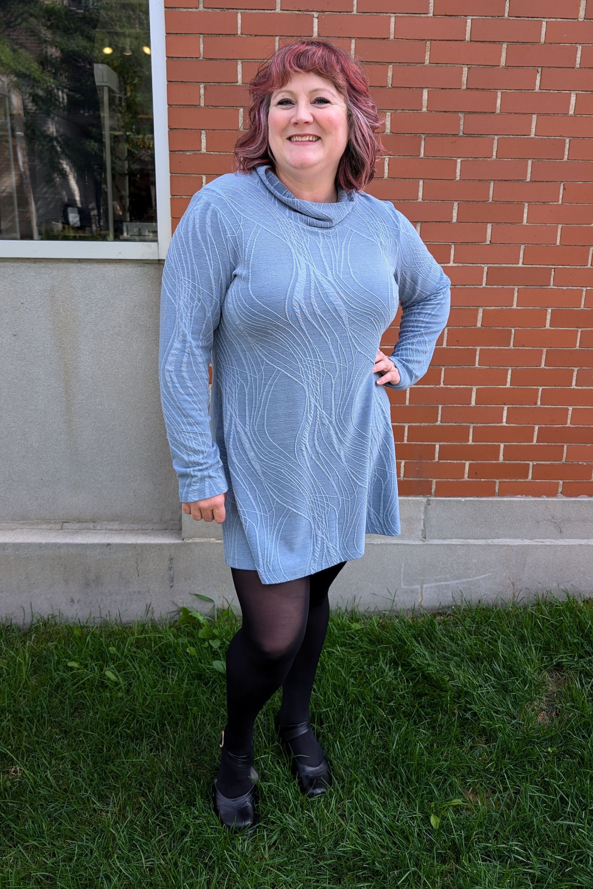 A woman wearing the Frisket Tunic by Luc Fontaine in Blue, featuring a textured fabric, cowl neck, long sleeves, and mid-thigh length, standing outside on the grass in front of a brick wall. 