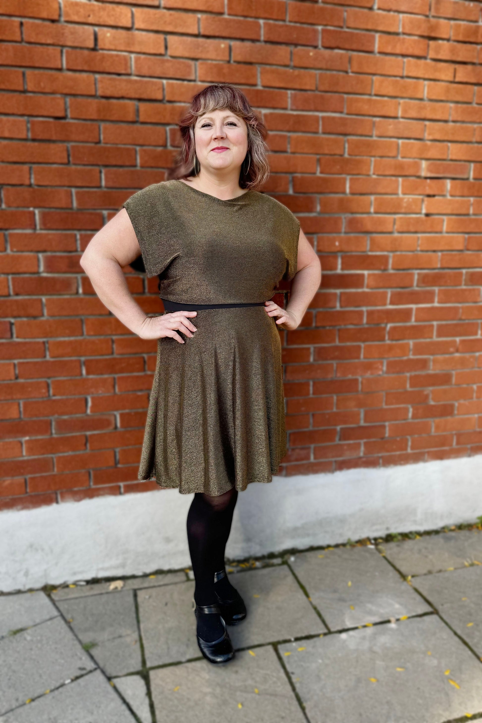 A woman wearing the Miyuki Dress by Infime in Gold, with glittery fabric, a boat neck, short extended sleeves, a contrast waistband, and a flared knee-length skirt, standing in front of a brick wall