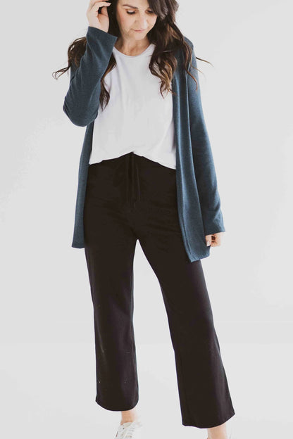A woman wearing the Carolina Cardigan by Ang Hill in Juniper, an open cardigan with long sleeves. She is wearing it with black pants and a white top and is standing in front of a white background. 
