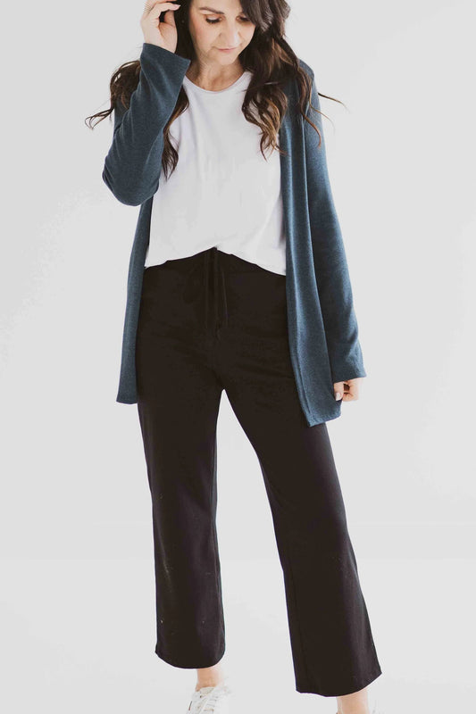 A woman wearing the Carolina Cardigan by Ang Hill in Juniper, an open cardigan with long sleeves. She is wearing it with black pants and a white top and is standing in front of a white background. 