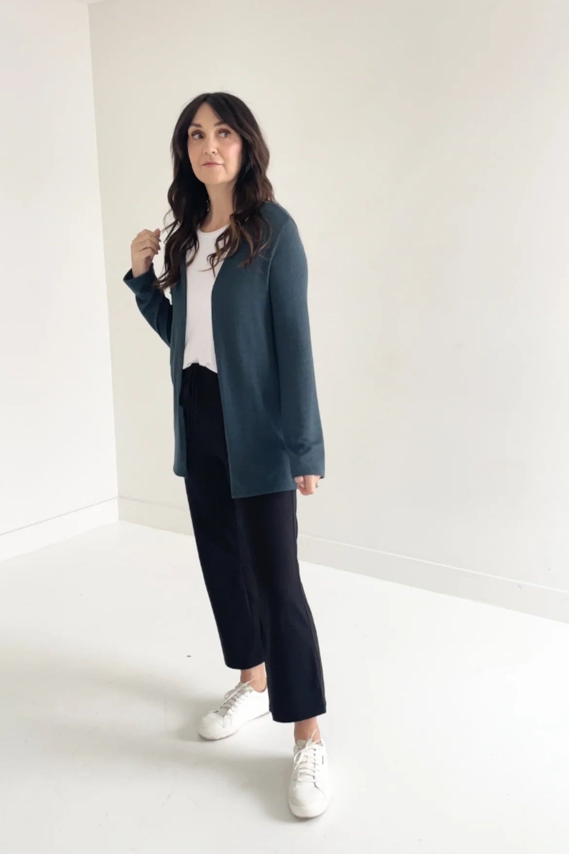 A woman wearing the Carolina Cardigan by Ang Hill in Juniper, an open cardigan with long sleeves. She is wearing it with black pants and a white top and is standing in front of a white background. 